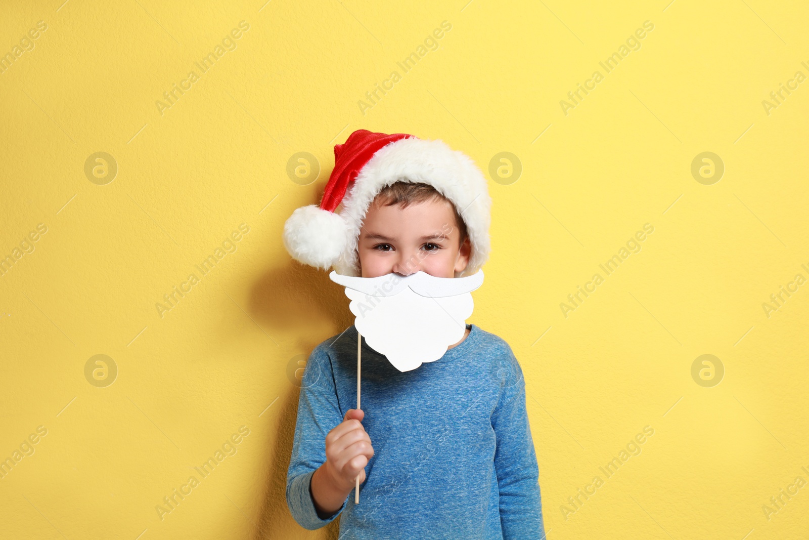 Image of Cute little boy with Santa hat and white beard prop on yellow background. Christmas celebration