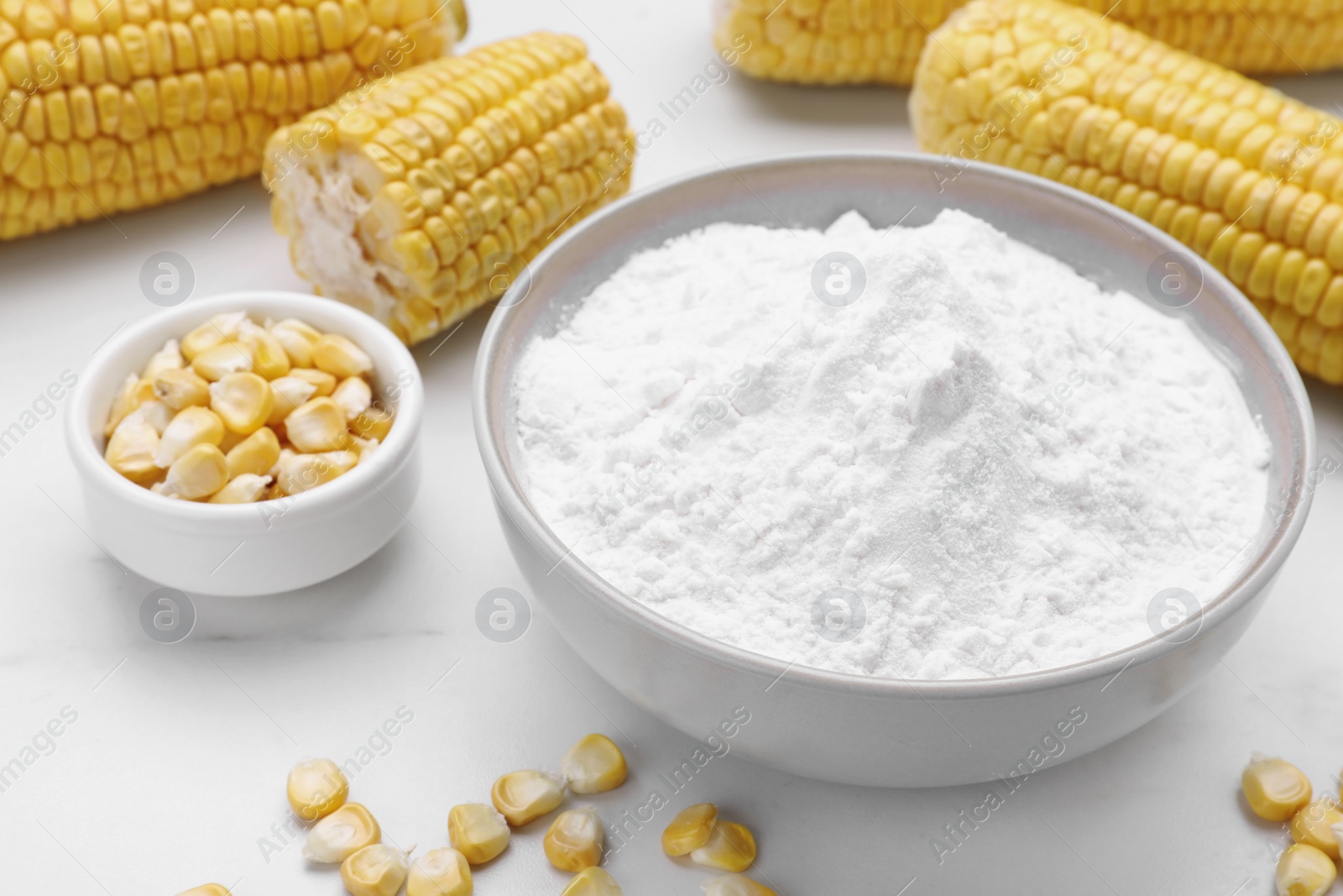 Photo of Bowl with corn starch, ripe cobs and kernels on white table, closeup
