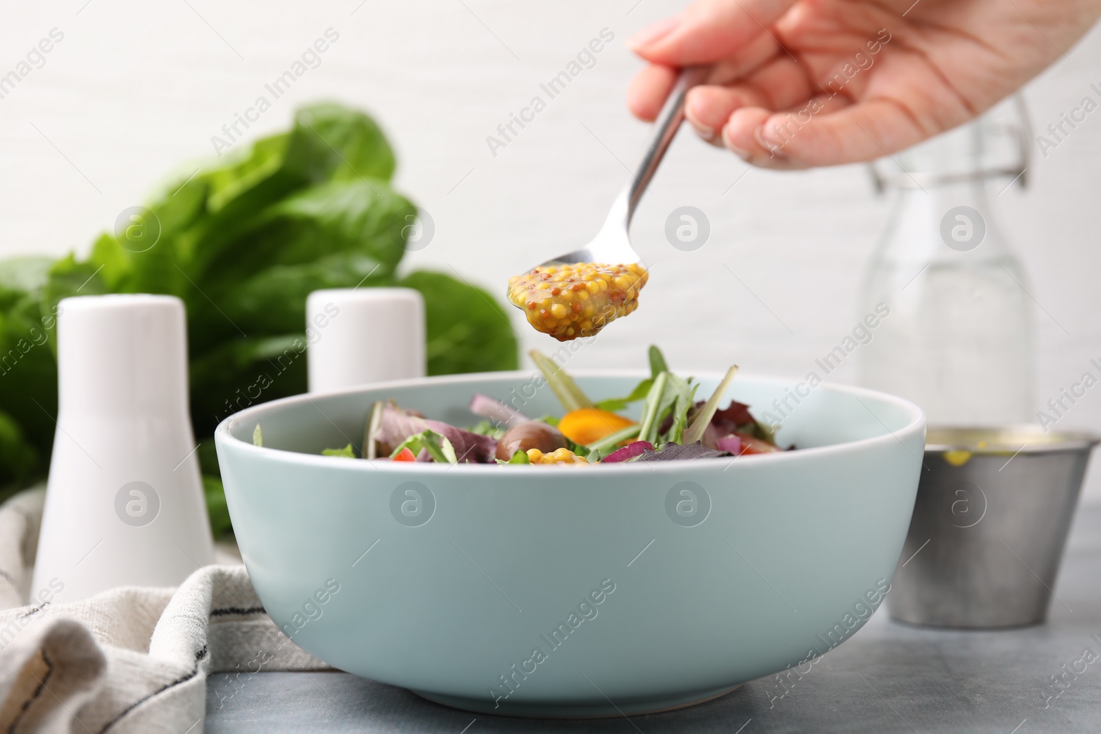 Photo of Woman pouring tasty vinegar based sauce (Vinaigrette) from spoon into bowl with salad at grey table, closeup