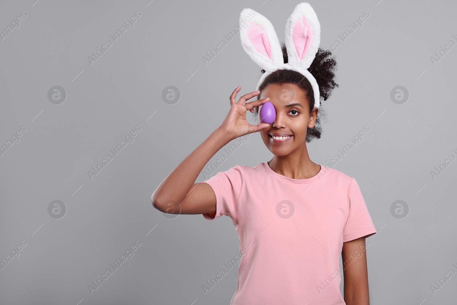 Photo of Happy African American woman in bunny ears headband covering eye with Easter egg on gray background. Space for text
