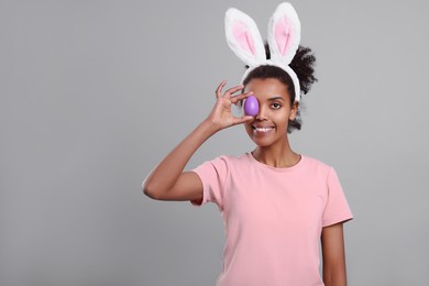 Happy African American woman in bunny ears headband covering eye with Easter egg on gray background. Space for text