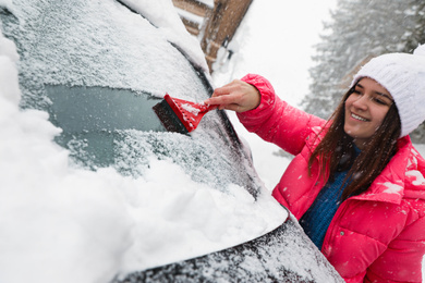 Photo of Young woman cleaning snow from car windshield outdoors on winter day