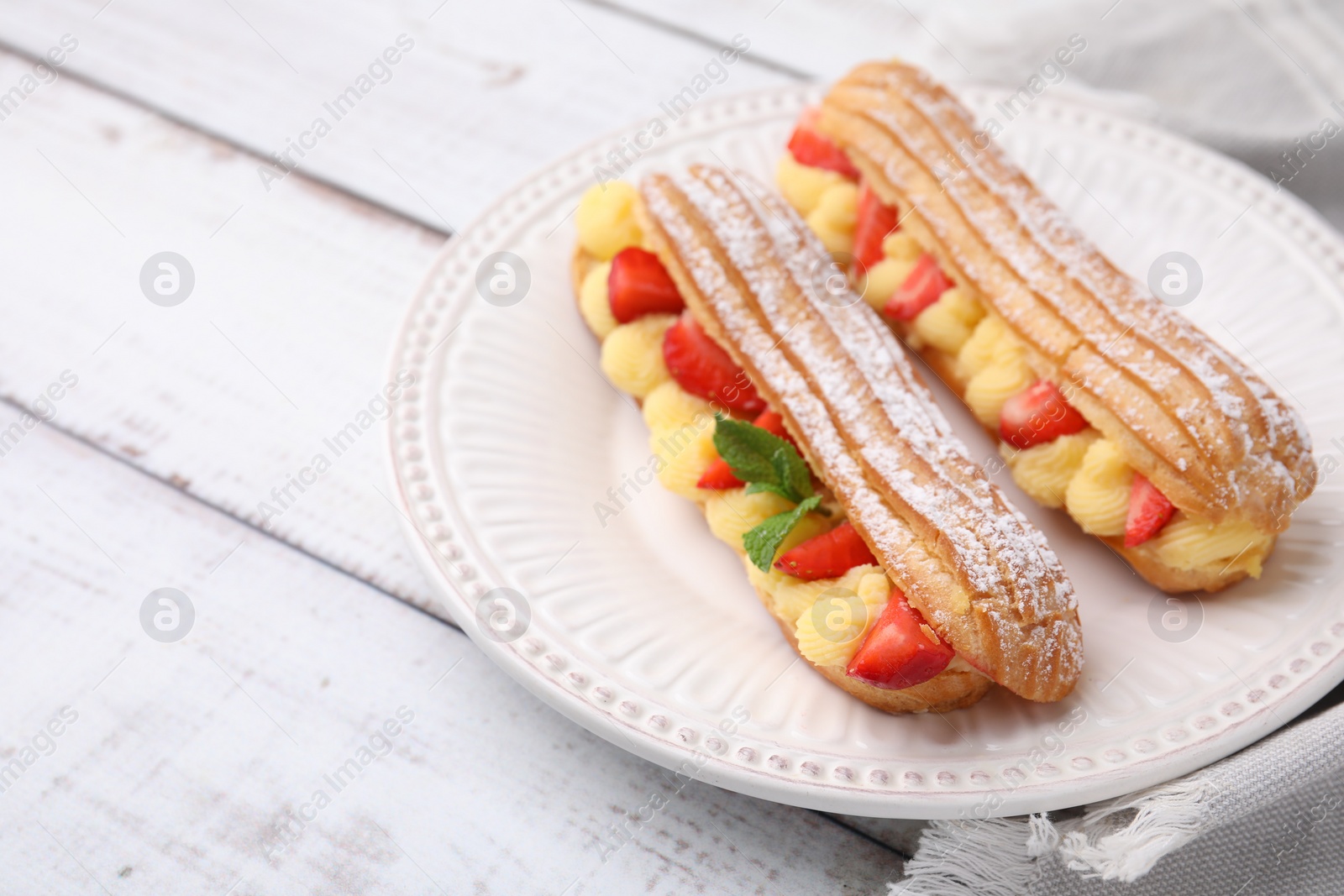 Photo of Delicious eclairs filled with cream, strawberries and mint on white wooden table