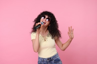Beautiful young woman with microphone and sunglasses singing on pink background