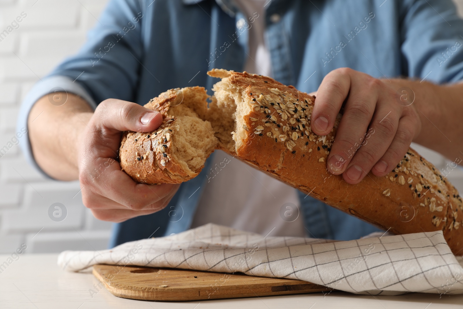 Photo of Man breaking loaf of fresh bread at white table near brick wall, closeup