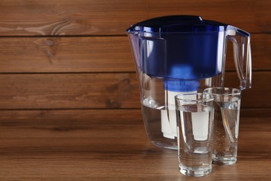 Photo of Filter jug and glasses with purified water on wooden table. Space for text
