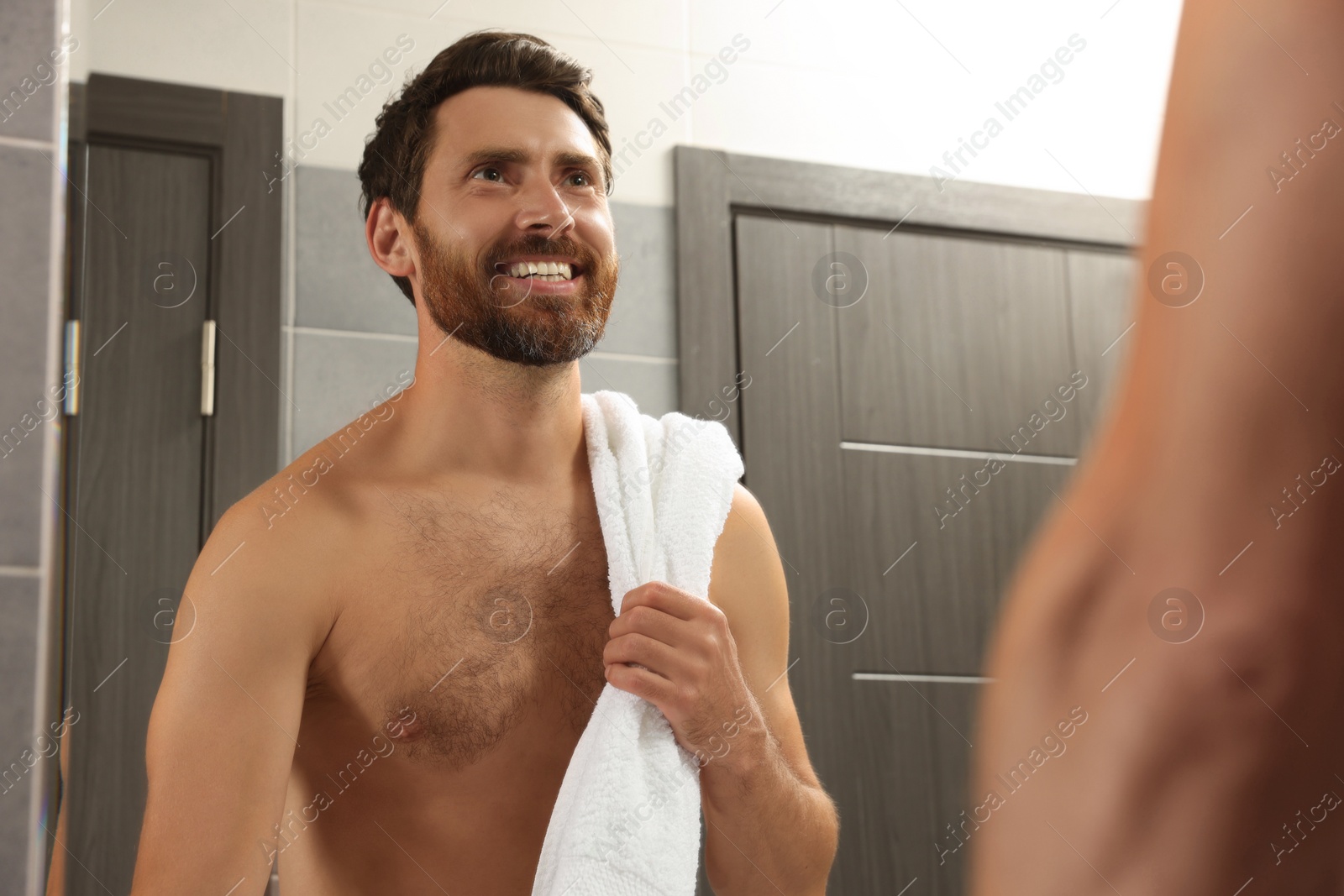 Photo of Handsome bearded man looking at mirror in bathroom near wooden doors