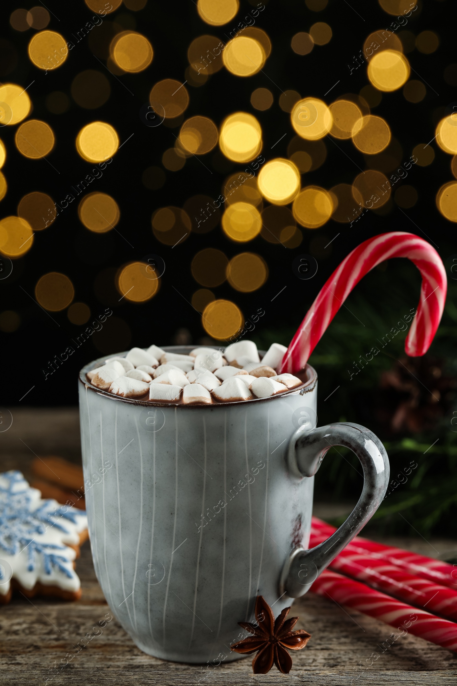 Photo of Delicious hot chocolate with marshmallows and candy cane on wooden table against blurred lights