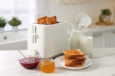 Photo of Making toasts for breakfast. Appliance, crunchy bread, honey, jam and milk on white marble table in kitchen