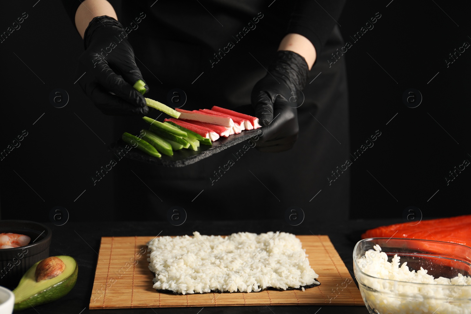 Photo of Chef in gloves putting cucumber onto unwrapped sushi roll at dark table, closeup