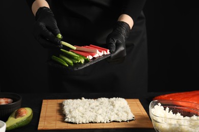 Chef in gloves putting cucumber onto unwrapped sushi roll at dark table, closeup