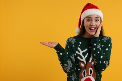 Photo of Happy young woman in Christmas sweater and Santa hat showing something on orange background. Space for text