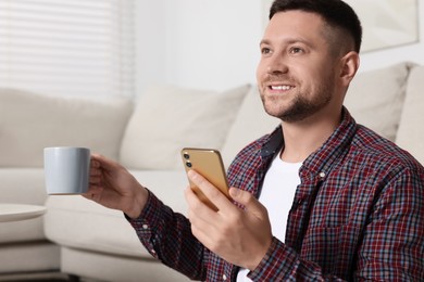 Handsome man with cup of drink sending message via smartphone at home