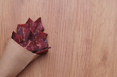 Paper bag with pieces of delicious beef jerky on wooden table, top view. Space for text
