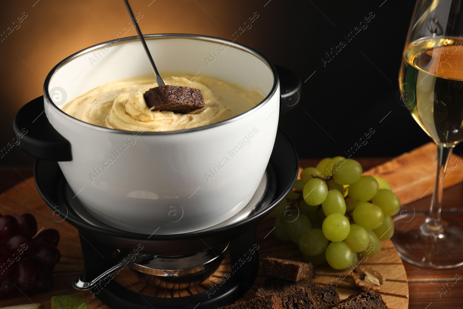 Photo of Dipping piece of bread into fondue pot with melted cheese at wooden table with snacks, closeup