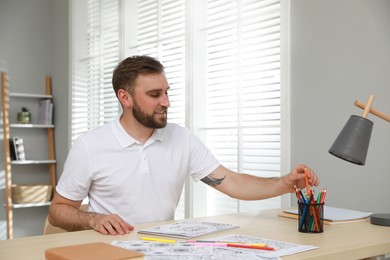 Young man coloring antistress picture at table indoors