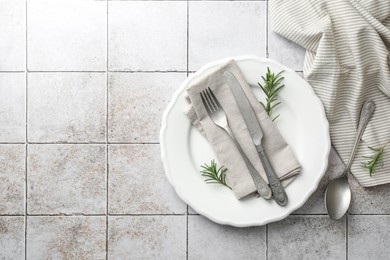 Stylish setting with cutlery, napkin, rosemary and plate on light tiled table, top view. Space for text