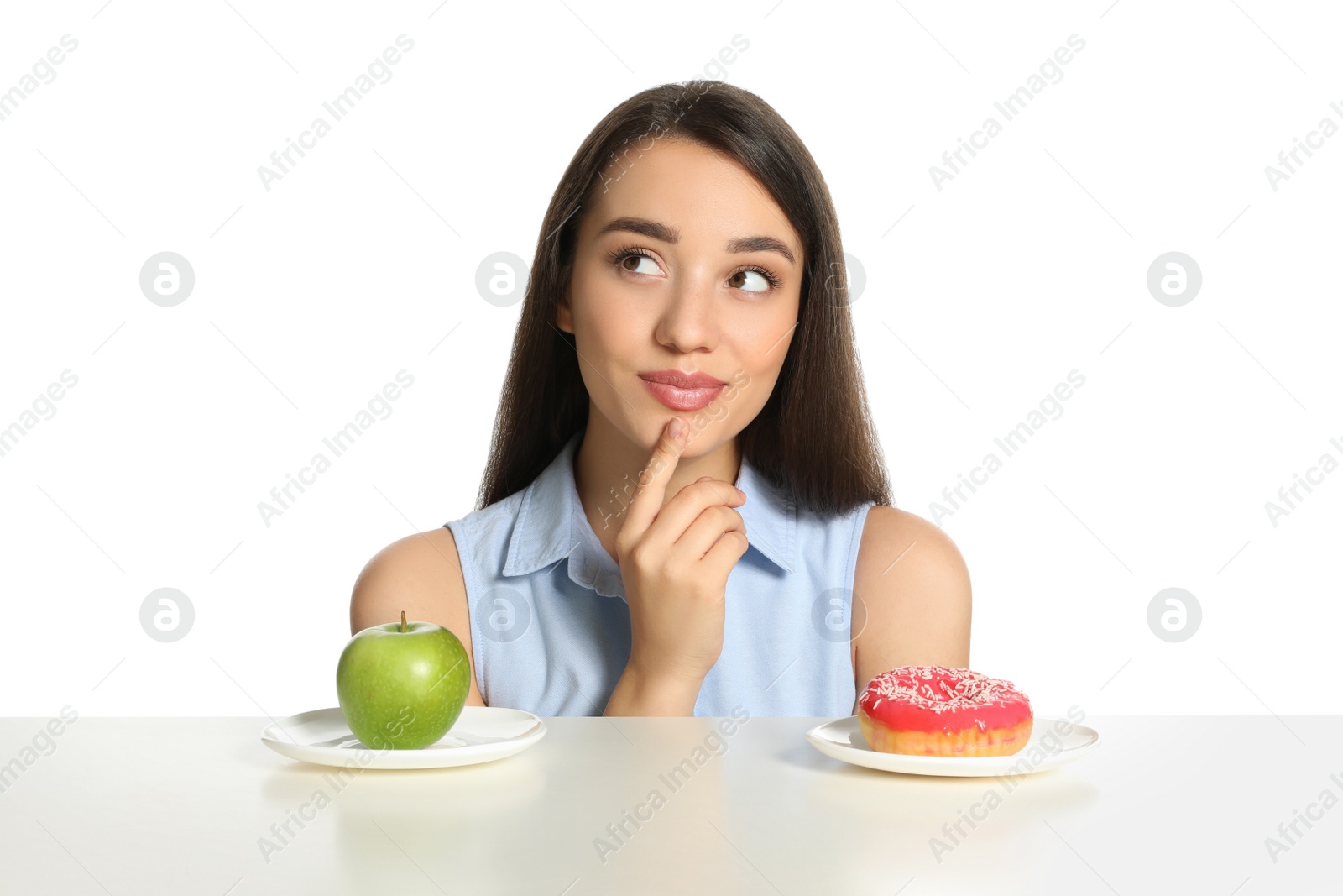 Photo of Doubtful woman choosing between apple and doughnut at table on white background