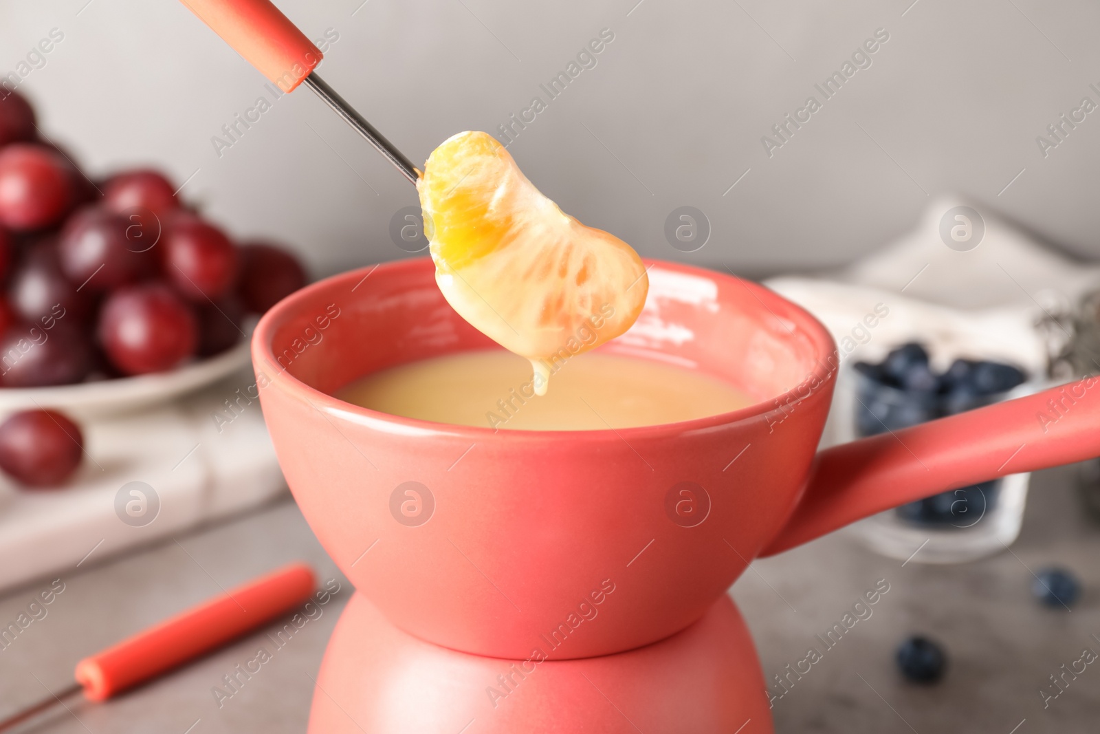 Photo of Dipping tangerine segment into pot with white chocolate fondue on table, closeup