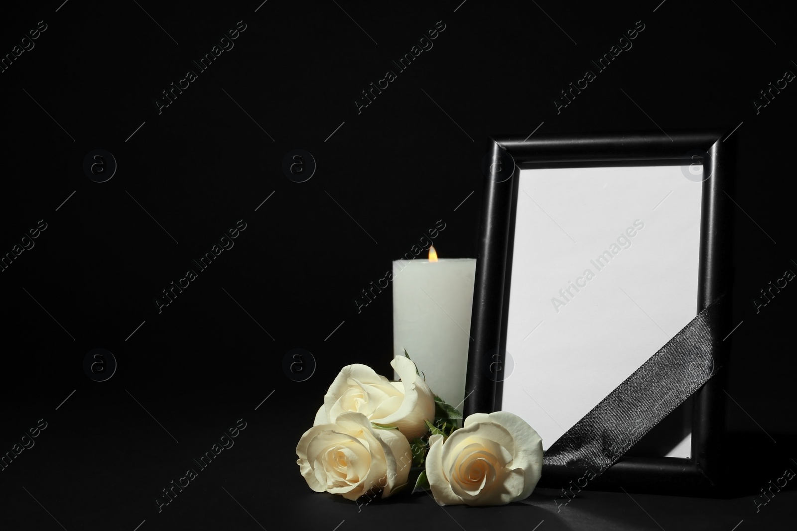 Photo of Funeral photo frame with ribbon, white roses and candle on dark table against black background. Space for design