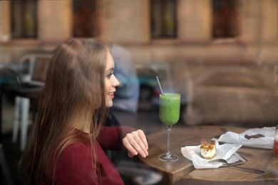 Photo of Pretty young woman with cocktail and cake at table in cafe, view from outdoors through window