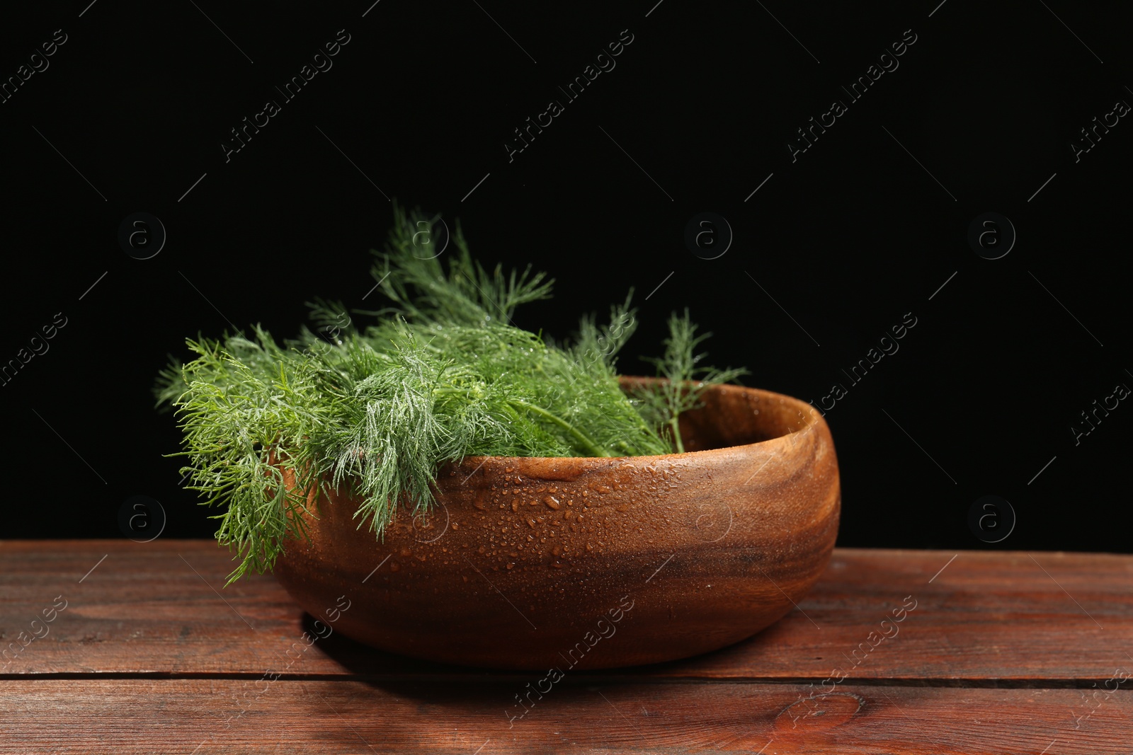 Photo of Bowl of fresh green dill with water drops on wooden table against black background