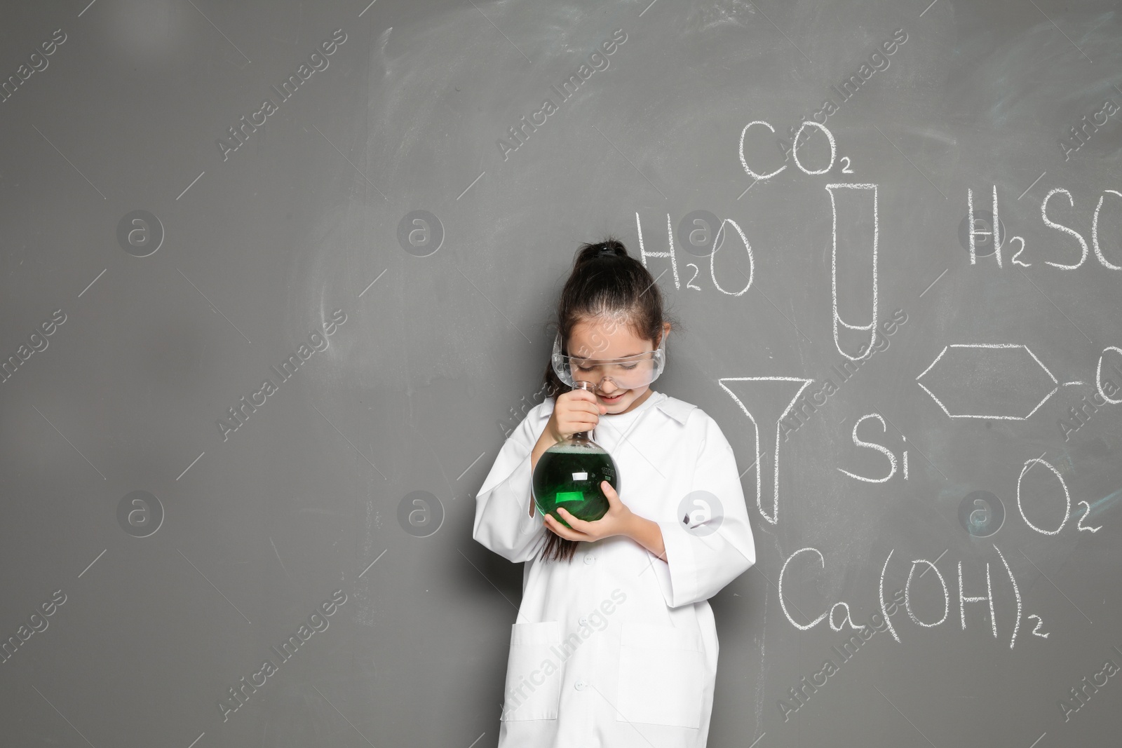 Photo of Little school child in laboratory uniform with flask of liquid and chemical formulas on grey background