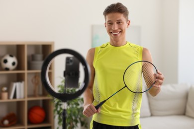 Smiling sports blogger holding badminton racket while streaming online fitness lesson with smartphone at home