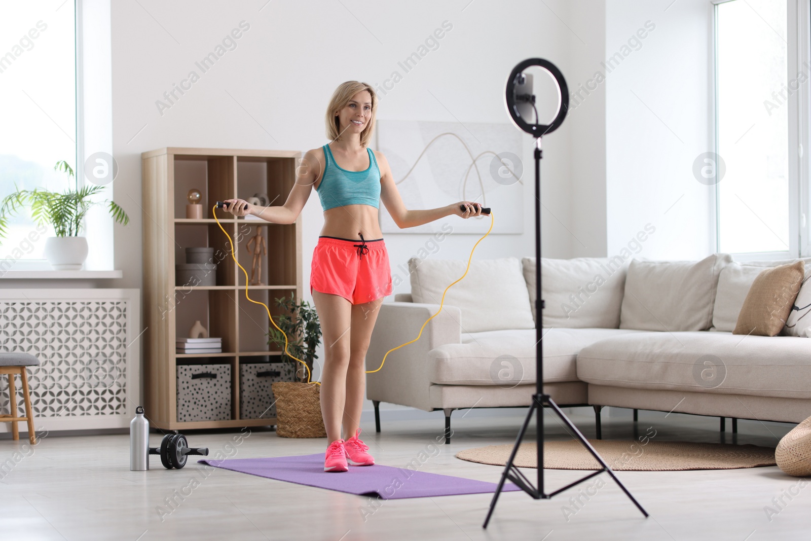 Photo of Smiling sports blogger jumping with rope while streaming online fitness lesson with smartphone at home