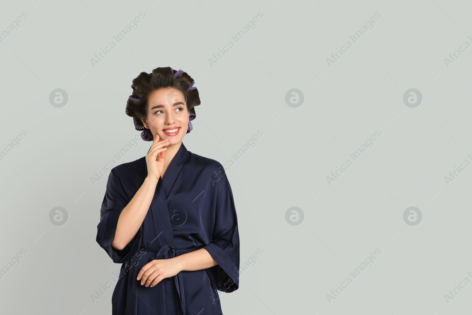 Photo of Happy young woman in silk bathrobe with hair curlers on grey background, space for text