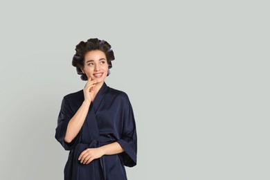 Photo of Happy young woman in silk bathrobe with hair curlers on grey background, space for text