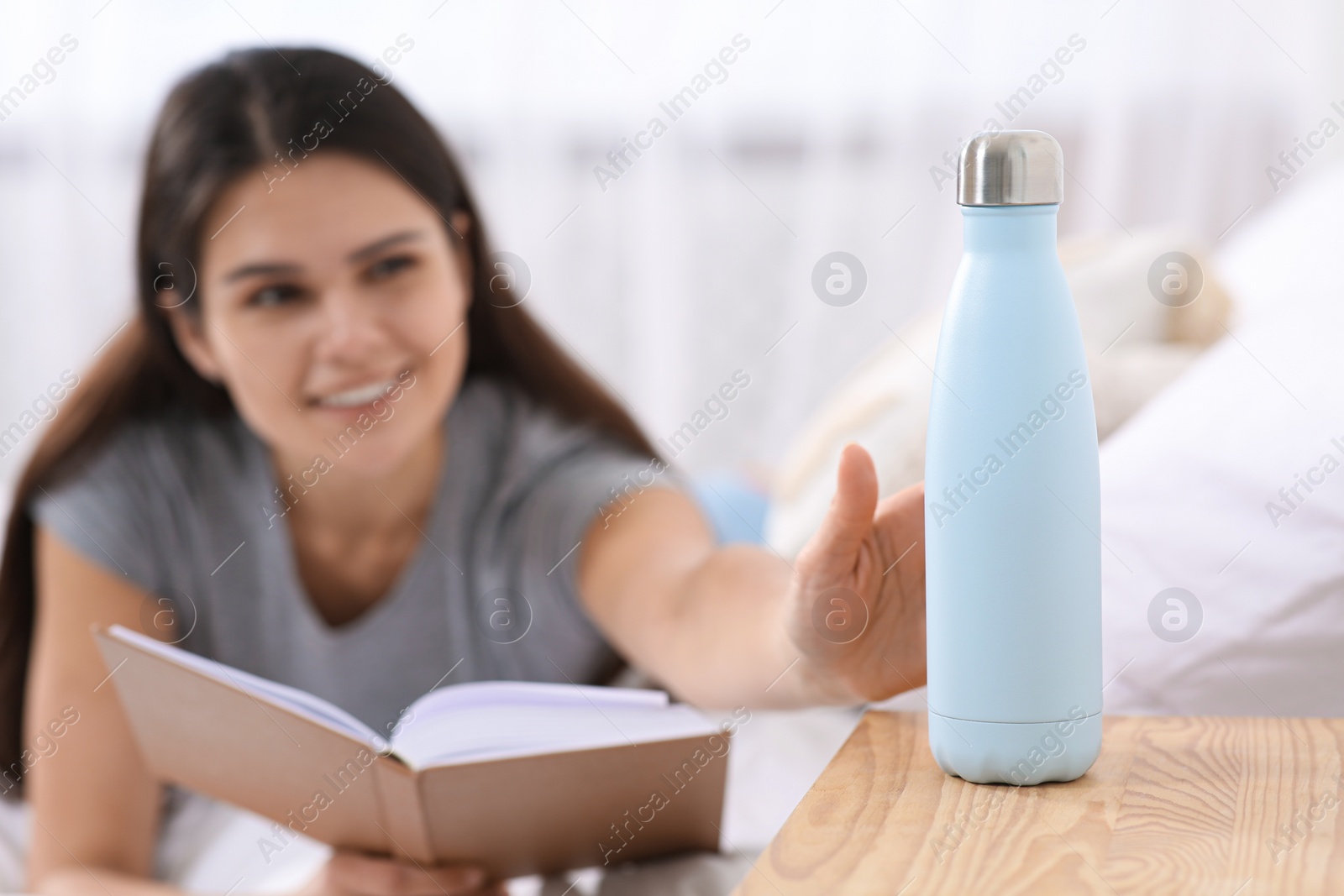 Photo of Young woman reading book in bedroom, focus on bottle
