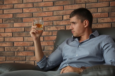 Young man with glass of whiskey near brick wall indoors