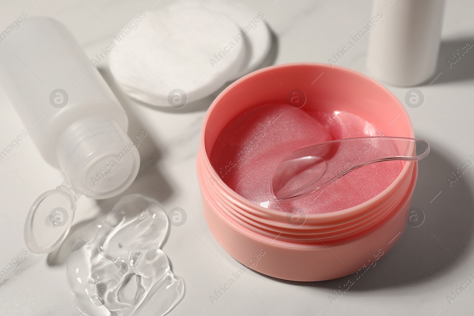 Photo of Jar of under eye patches with spoon and cosmetic products on white marble table, closeup