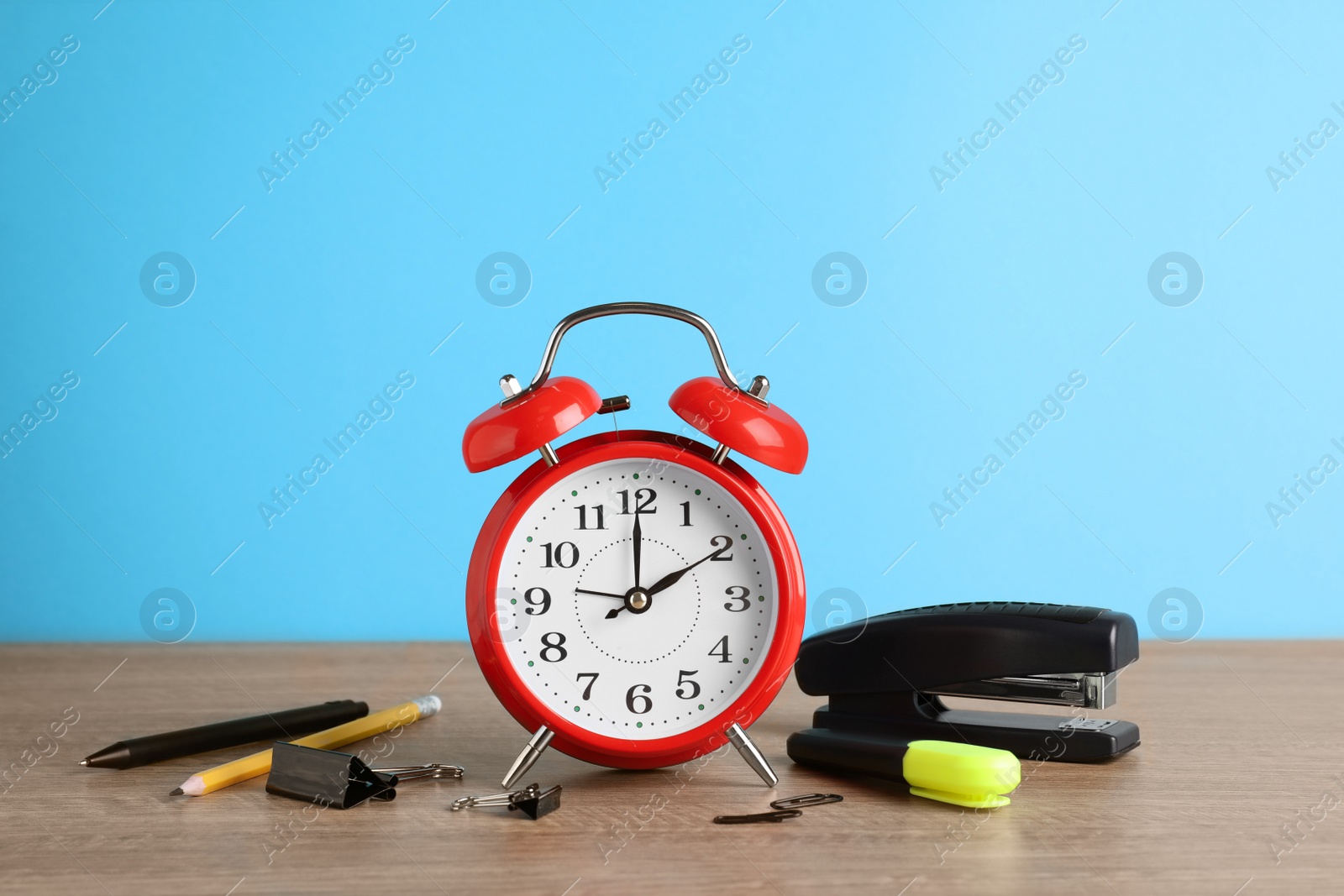 Photo of Red alarm clock and different stationery on wooden table against light blue background. School time