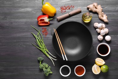 Photo of Empty iron wok and chopsticks surrounded by ingredients on dark grey wooden table, flat lay. Space for text