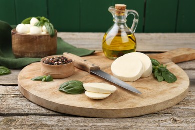 Photo of Board with tasty mozzarella slices, basil leaves, knife and spices on wooden table