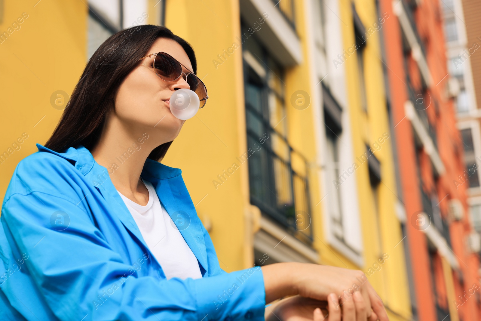 Photo of Beautiful young woman with sunglasses blowing chewing gum on city street, low angle view