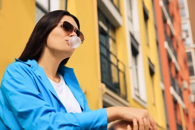 Photo of Beautiful young woman with sunglasses blowing chewing gum on city street, low angle view