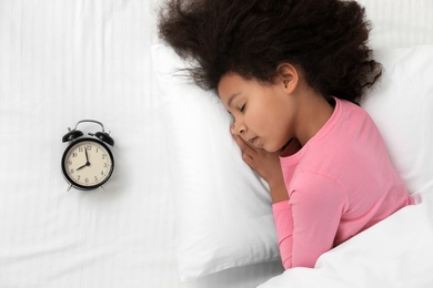 Photo of Cute little African-American girl with alarm clock sleeping in bed, top view