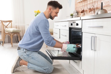 Photo of Handsome man taking out tray of baked cookies from oven in kitchen