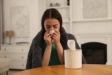 Photo of Sick woman wrapped in blanket with tissue blowing nose at wooden table indoors. Cold symptoms