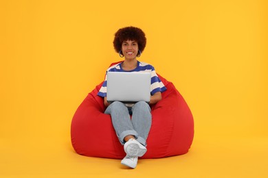 Beautiful young woman with laptop sitting on beanbag chair against orange background