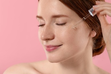 Beautiful woman with freckles applying cosmetic serum onto her face on pink background, closeup