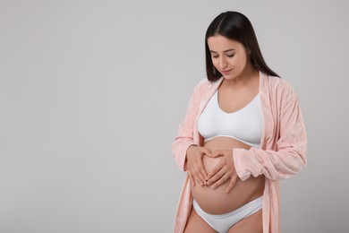 Photo of Beautiful pregnant woman in stylish comfortable underwear and robe making heart with hands on her belly against grey background, space for text