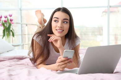 Photo of Attractive young woman using mobile phone and laptop on bed at home