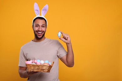 Photo of Happy African American man in bunny ears headband holding wicker tray with Easter eggs on orange background, space for text