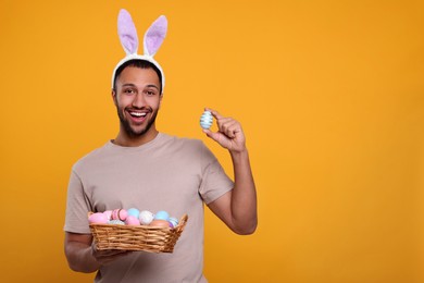 Happy African American man in bunny ears headband holding wicker tray with Easter eggs on orange background, space for text