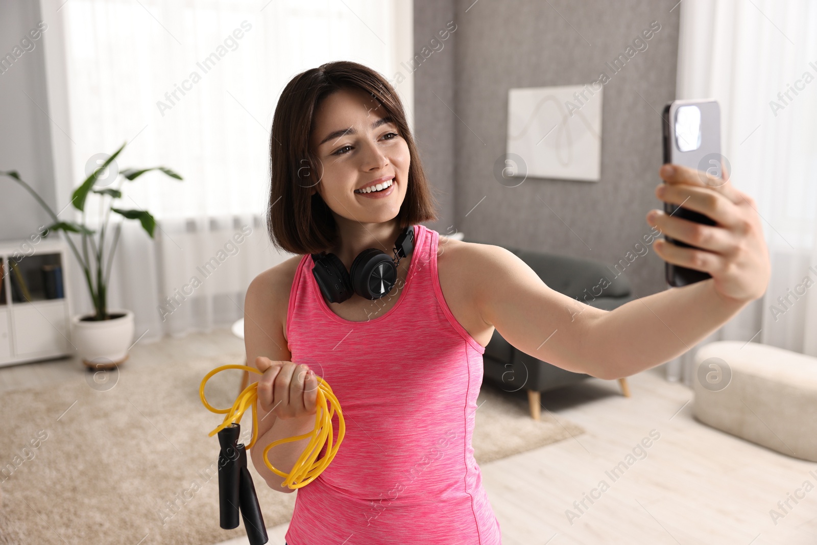 Photo of Happy sports blogger holding skipping rope while streaming online fitness lesson with smartphone at home