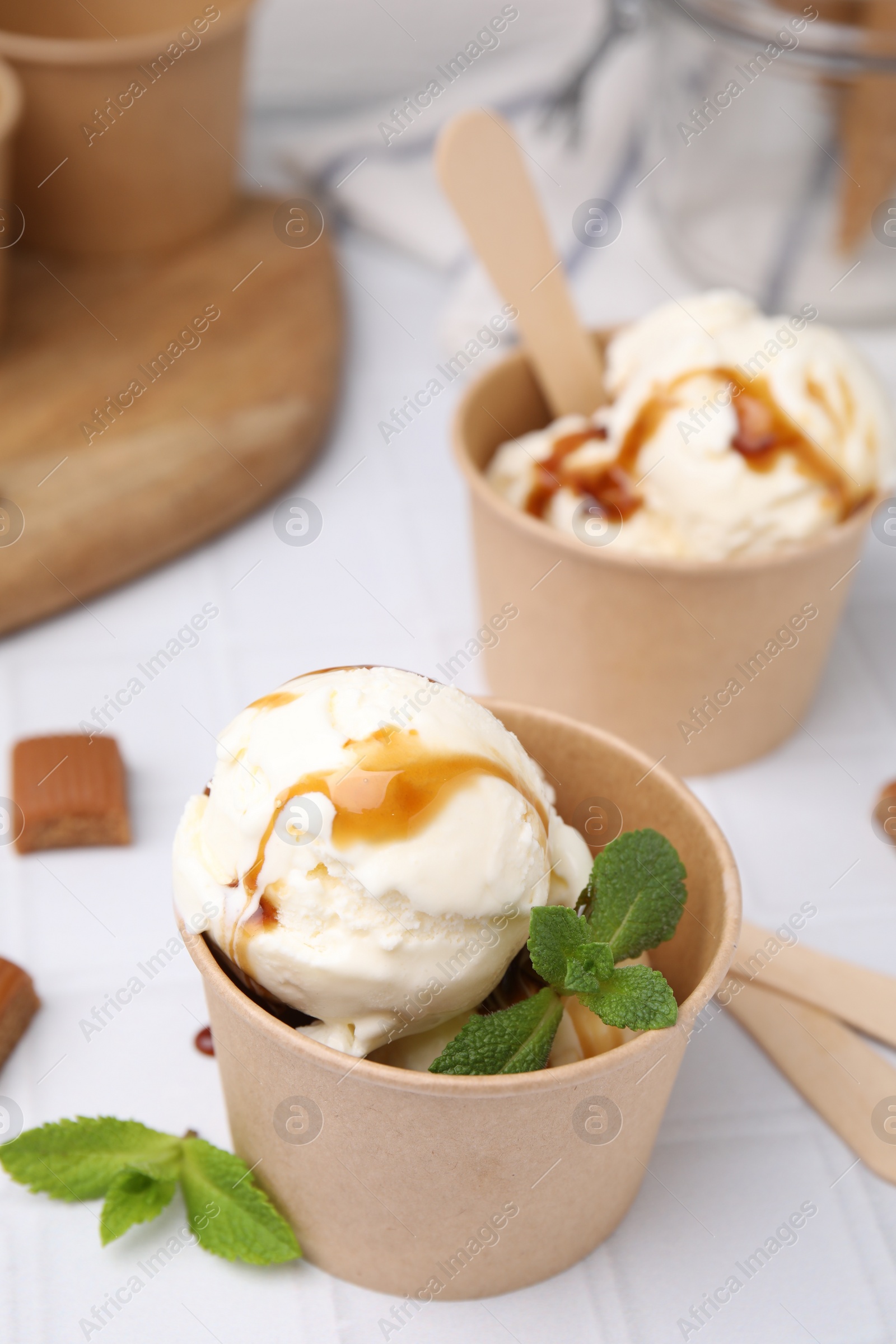 Photo of Scoops of ice cream with caramel sauce, mint leaves and candies on white tiled table, closeup